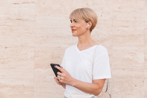 Foto de una mujer rubia europea con camiseta blanca que usa el teléfono móvil para escuchar música, mientras está de pie contra la pared beige al aire libre con auricular bluetooth