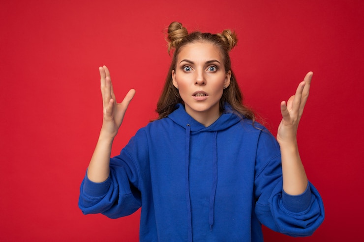 Foto foto de mujer rubia hermosa joven emocional sorprendida con dos cuernos con emociones sinceras con sudadera con capucha azul brillante hipster aislada sobre fondo rojo con espacio libre. wow concepto.
