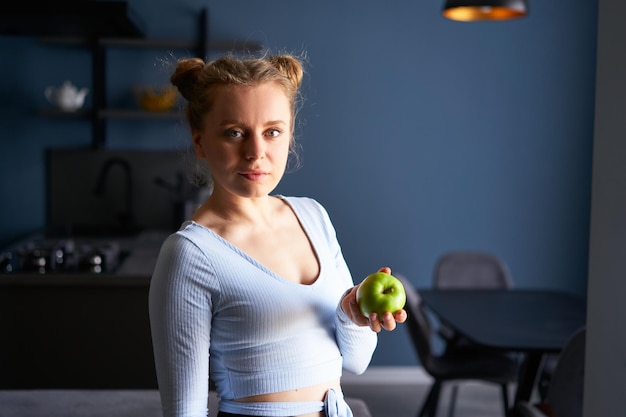 Foto de mujer sana y encantadora joven caucásica tranquila con maquillaje natural sosteniendo manzana verde sobre la oscuridad