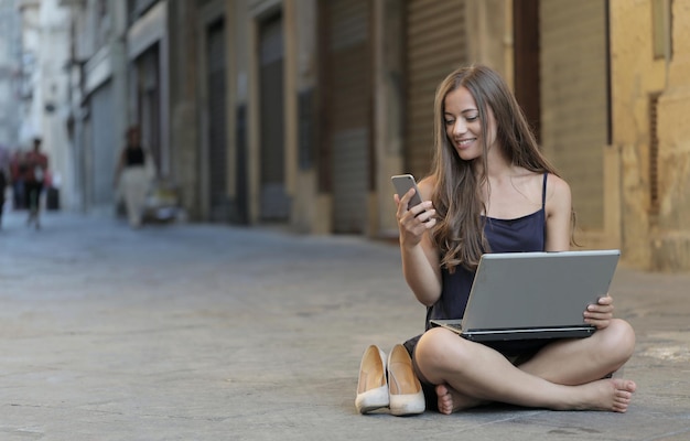 Foto de una mujer sentada en el suelo mientras usa un teléfono inteligente y una computadora portátil