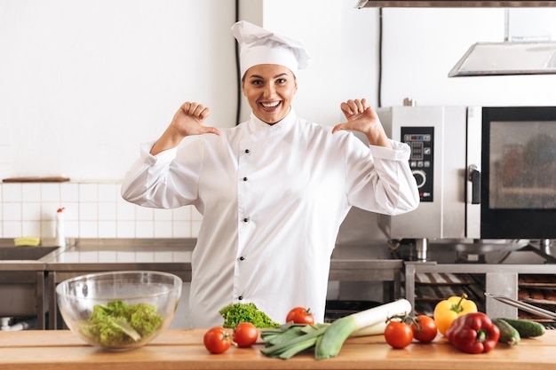 Foto de mujer sonriente chef vistiendo uniforme blanco cocinar comida con verduras frescas, en la cocina del restaurante