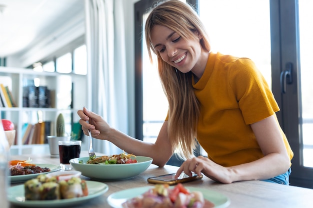 Foto de mujer sonriente comiendo alimentos saludables mientras usa su teléfono móvil en casa.