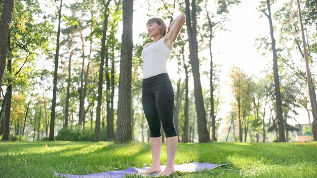 Foto de mujer sonriente de mediana edad practicando yoga y meditando en el parque. Mujer estirando y haciendo fitness en mat en el bosque