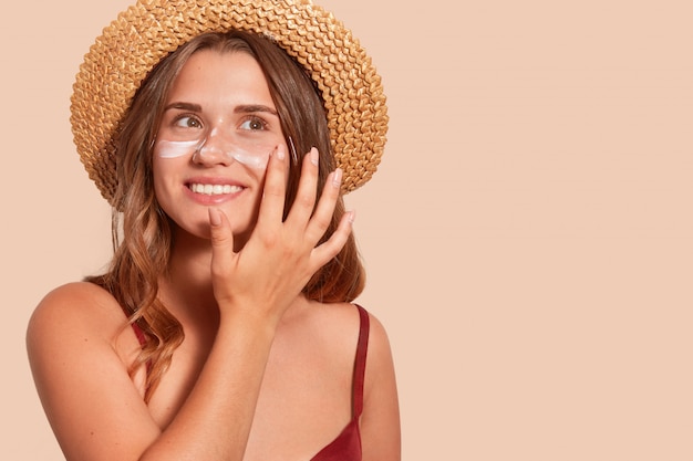 Foto de mujer sonriente con el pelo largo, tiene feliz expresión facial, aplaudiendo protector solar, con sombrero de paja, queriendo broncearse, aislado en la pared de color beige. Verano, vacaciones, concepto de protector solar.