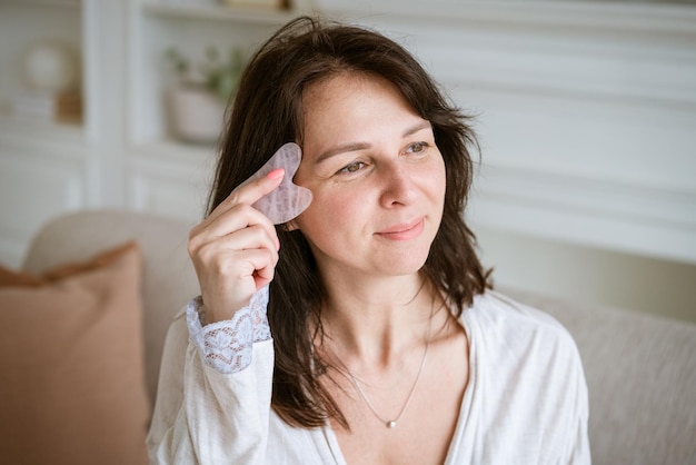 Una foto de una mujer sosteniendo un raspador de gouache rosa para masajear su rostro durante su rutina de belleza matutina en
