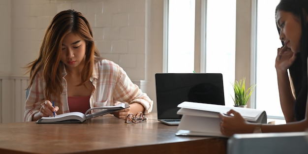 Foto de mujeres hermosas que leen un libro y se relajan juntas mientras que se sientan en la mesa de madera sobre sala de estar cómoda. Mujeres con concepto de tiempo relajante.