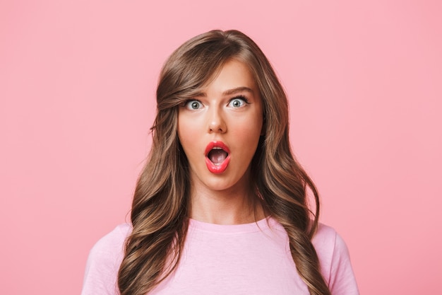 Foto-Nahaufnahme der aufgeregten hübschen Frau mit dem langen lockigen Haar im einfachen T-Shirt, das Überraschung mit offenem Mund und prall gefüllten Augen ausdrückt, lokalisiert über rosa Hintergrund