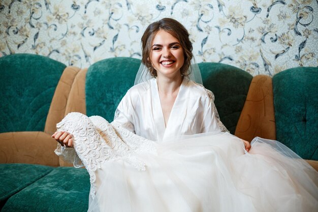 Foto Nahaufnahme Porträt einer Braut mit professionellem Make-up ist sie in einem weißen Mantel Schleier sitzt auf der Couch und Blick auf das Hochzeitskleid Junge Frau Hochzeitstag Foto-Shooting Frisur Schönheit