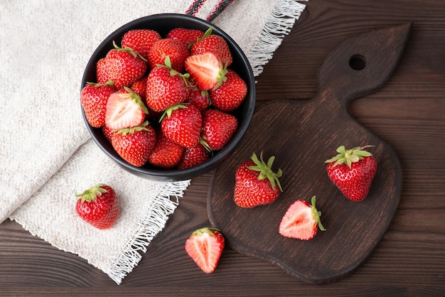 Foto de naturaleza muerta de fresas en un tazón negro, tabla de cortar de madera y toalla blanca hecha de lino natural Sabrosa fresa fresca