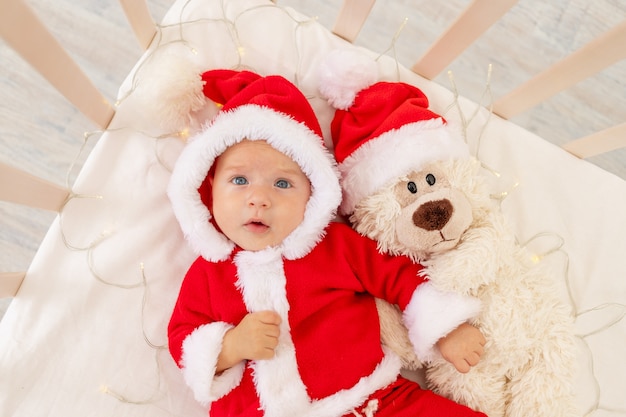 Foto de Navidad de un bebé con un disfraz de Papá Noel acostado en una cuna en casa con un juguete en un gorro de Papá Noel