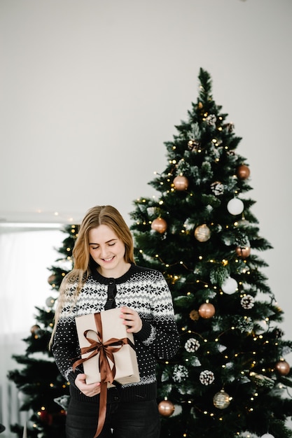 Foto de Navidad de niña con cabello rubio en ropa acogedora posando cerca del árbol de Navidad con caja de regalos