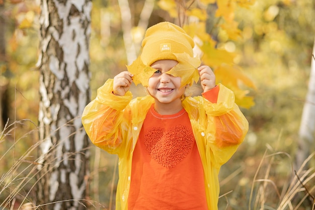 Foto de niña de 5 años feliz de disfrutar en otoño, se puso un sombrero amarillo y un impermeable, cerró los ojos con hojas otoñales de árboles.