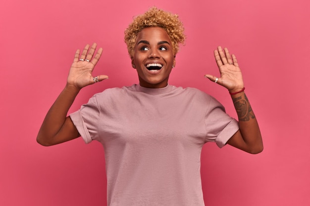 La foto de una niña afroamericana baila con suerte, levanta las manos en señal de alegría, se siente campeona después de obtener el triunfo, mira alegremente a algún lado, se divierte, siente el ritmo de la música con un fondo rosa