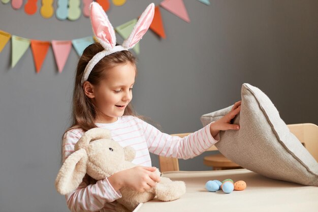 Foto de una niña alegre y sonriente con orejas de conejo encuentra huevos debajo de la almohada Caza de Pascua posando en el interior de la casa con decoraciones celebrando las fiestas jugando