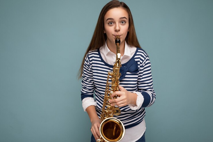Foto foto de niña brunet feliz bastante positiva vistiendo elegante manga larga a rayas que se encuentran aisladas sobre fondo azul pared tocando un instrumento musical mirando a la cámara