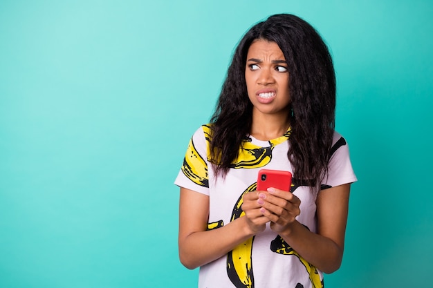 Foto de niña disgustada disgustada mantenga teléfono mire espacio vacío use camiseta con estampado de plátano aislado de fondo de color turquesa