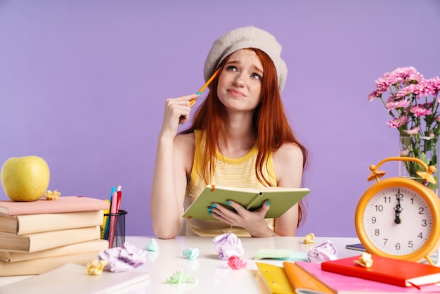 Foto de niña estudiante frustrada escribiendo en el libro de ejercicios mientras hace la tarea aislada sobre la pared púrpura