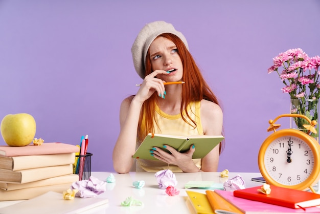 Foto foto de niña estudiante reflexiva escribiendo en el libro de ejercicios mientras hace la tarea aislada sobre la pared púrpura