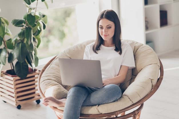 Foto de niña inteligente sentarse sillón de mimbre trabajo portátil llevar camiseta blanca jeans en casa en interiores