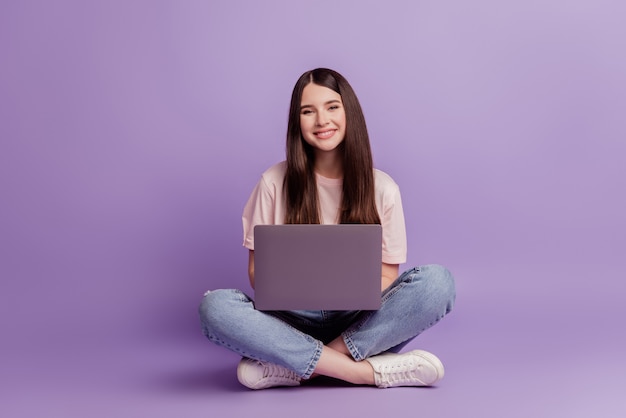 Foto de niña con laptop sentarse en el piso mirar la cámara sobre fondo púrpura