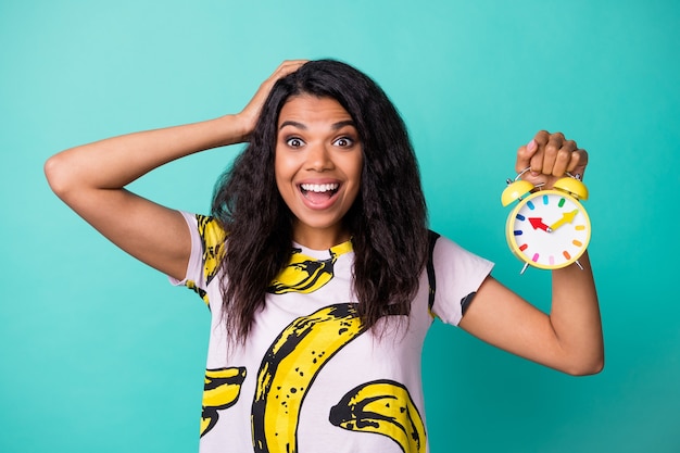 Foto de niña mantenga la mano del reloj despertador con la cabeza abierta con la boca abierta use camiseta con estampado de plátano aislado de fondo de color verde azulado