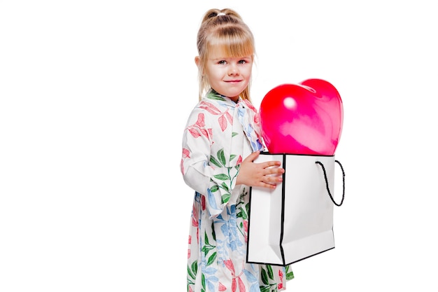 La foto de la niña de moda con un vestido de flores blanco sostiene una bolsa grande con un globo en forma de corazón en el interior.