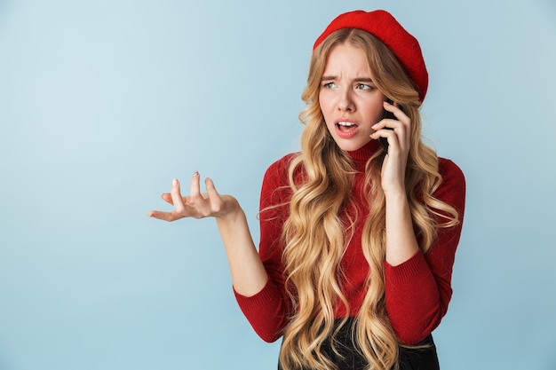 Foto de niña molesta de 20 años con boina roja hablando por teléfono inteligente aislado