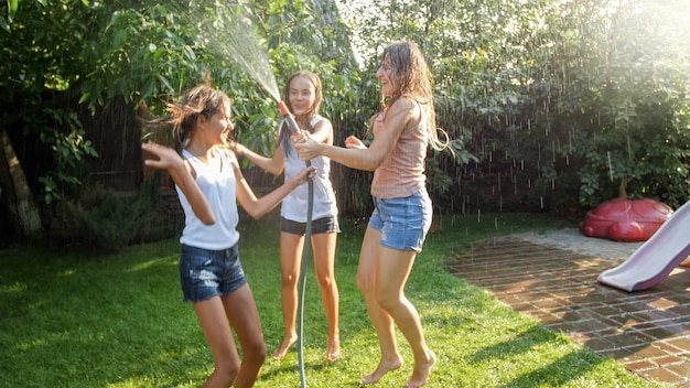 Foto de niñas alegres felices en ropa mojada bailando y saltando bajo la manguera del jardín de agua. Familia jugando y divirtiéndose al aire libre en verano