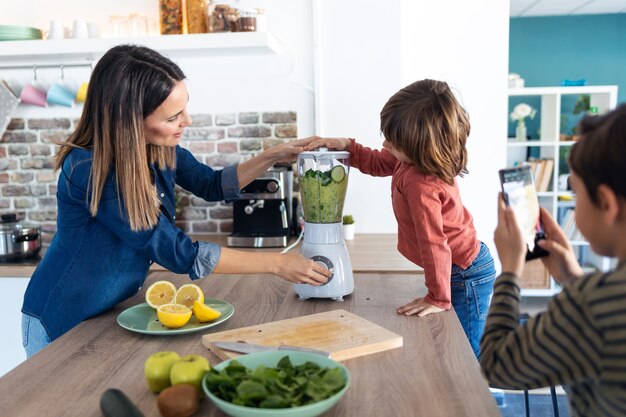 Foto de niño ayudando a su madre a preparar un jugo detox con licuadora mientras su hermano toma fotografías con el teléfono móvil en la cocina de su casa.