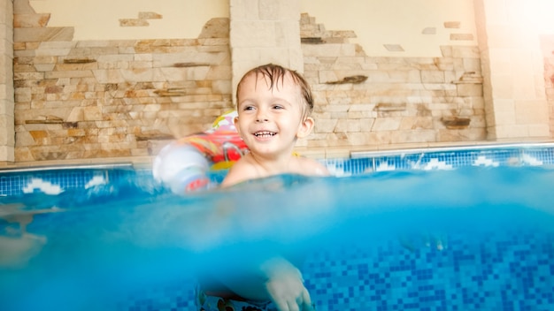 Foto de niño feliz riendo y sonriente jugando con juguetes y aprendiendo a nadar en la piscina interior