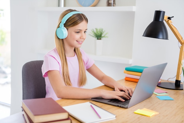 Foto de niño pequeño, niña, sentarse cómodamente, mesa acogedora, escritorio, estudio, uso remoto, computadora portátil, tener videollamadas en línea, comunicación con el maestro, enviar mensajes de texto, escribir, tener auriculares en casa en el interior