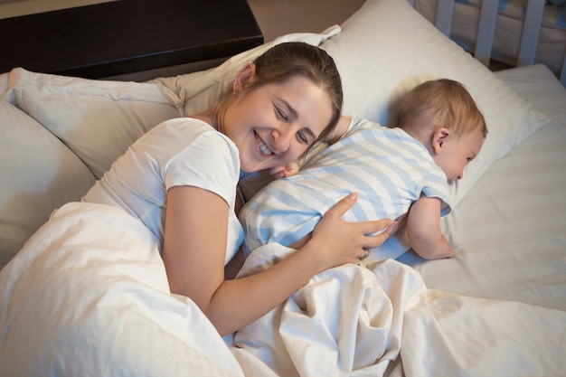 Foto nocturna de feliz madre sonriente acostada en la cama con su pequeño bebé