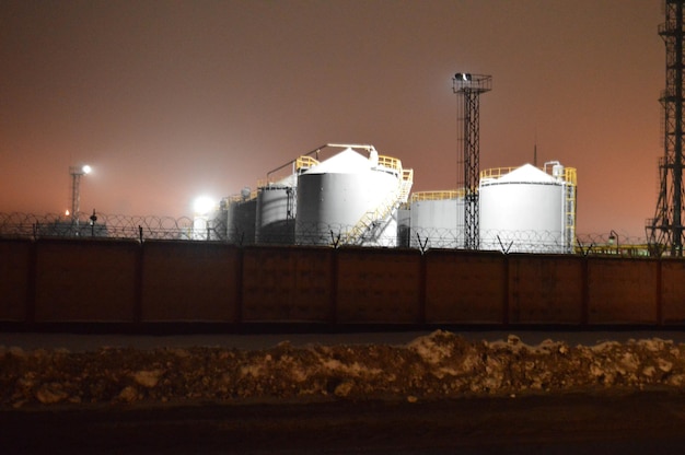 Foto noturna de uma fazenda de tanques de uma refinaria de petróleo, uma planta petroquímica através de uma cerca