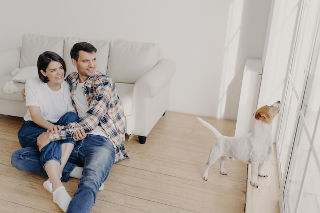 Una foto de un novio y una novia felices miran felizmente a su mascota que está cerca del balcón quiere jugar afuera pasar el fin de semana en casa retirarse en un apartamento recién comprado tener relaciones perfectas