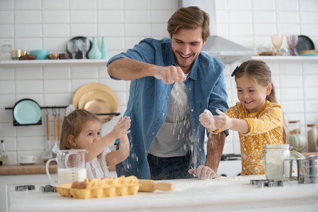 Foto de un padre sonriente y sus hijas horneando en la cocina y divirtiéndose.