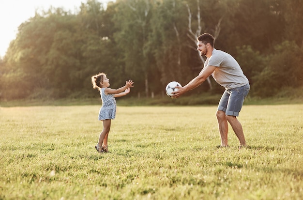 Foto de papá con su hija lanzando una pelota al hermoso césped y bosques al fondo.