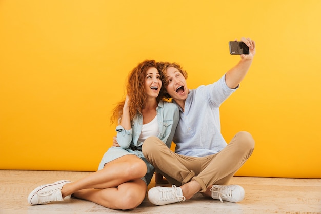 Foto de pareja alegre hombre guapo y mujer rizada sentados juntos en el suelo y tomando selfie en smartphone, aislado sobre fondo amarillo