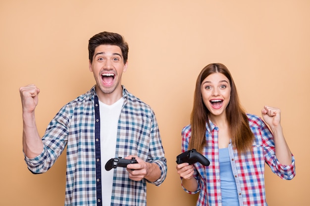 Foto de pareja alegre positiva casual de pelo marrón blanco jugando videojuegos de playstation regocijándose con la victoria sosteniendo joysticks con las manos aisladas sobre fondo de color beige pastel