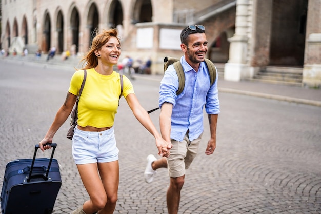 Foto de pareja joven cogidos de la mano y corriendo en la ciudad con maleta en vacaciones.