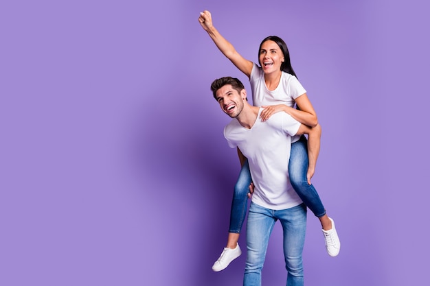 Foto de una pareja muy linda a cuestas en jeans denim camiseta blanca con el hombre que lleva a la niña dirigiéndolo hacia adelante sonriendo con dientes aislados de fondo de color pastel violeta