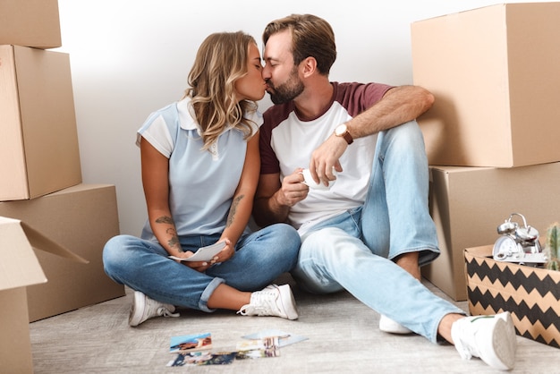 Foto de una pareja romántica en ropa casual tomando café y besándose mientras está sentado cerca de cajas de cartón aisladas sobre una pared blanca