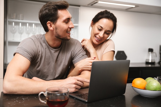 Foto de pareja sonriente hombre y mujer sentados en la cocina y usando la computadora portátil, mientras desayunan juntos