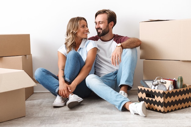 Foto de pareja sonriente en ropa casual abrazándose mientras está sentado cerca de cajas de cartón aisladas sobre pared blanca