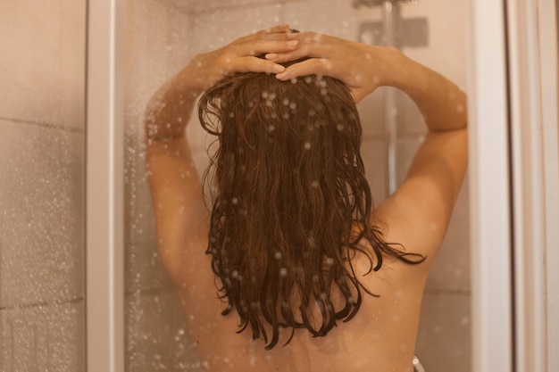 Foto foto de la parte posterior de la mujer de cabello oscuro con cuerpo desnudo se ducha detrás de la puerta de vidrio en el baño, disfruta de la relajación en el baño, tiene una piel sana y pura.