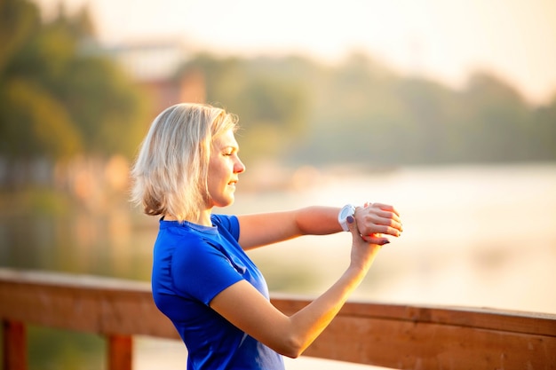 Foto de la parte posterior de la mujer deportiva mira el reloj en la mano, de pie sobre el puente de madera en el parque el día de verano