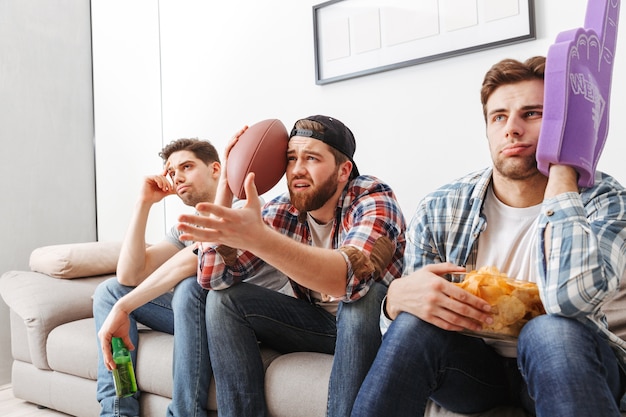 Foto de partidarios masculinos sin afeitar que expresan tristeza, mientras ven un juego deportivo en casa y apoyan al equipo de fútbol