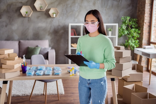 Foto de pedidos organizados de negocios de dama asiática, paquetes de máscaras médicas para la gripe facial para entregar cajas en espera, servicio de correos, retención del portapapeles, verificación de números de información, oficina en casa en el interior