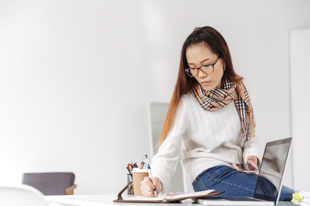 Foto de pensamiento trabajadora asiática con gafas usando el teléfono celular y escribiendo en su diario mientras está sentado a la mesa en la oficina