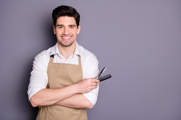 Foto de perfil de atractivo trabajador chico mantenga peinado herramientas de corte tijeras peine estilista de cabello profesional invitando al cliente sentarse usar delantal camisa blanca aislada pared de color gris