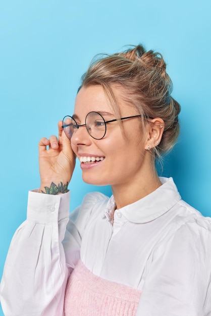 Foto de perfil de colegiala feliz mantiene la mano en el borde de las gafas sonríe con alegría viste camisa blanca nota algo divertido en la distancia aislada sobre fondo azul. Mujer encantada complacida